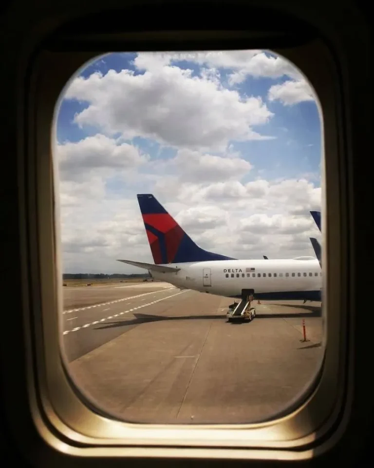 View through an airplane window of a Delta jet parked on the tarmac under a partly cloudy sky.