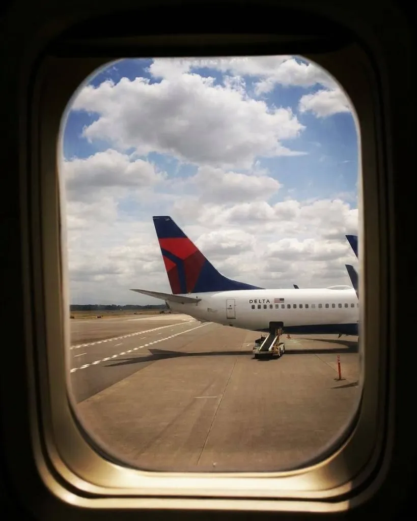 View through an airplane window of a Delta jet parked on the tarmac under a partly cloudy sky.